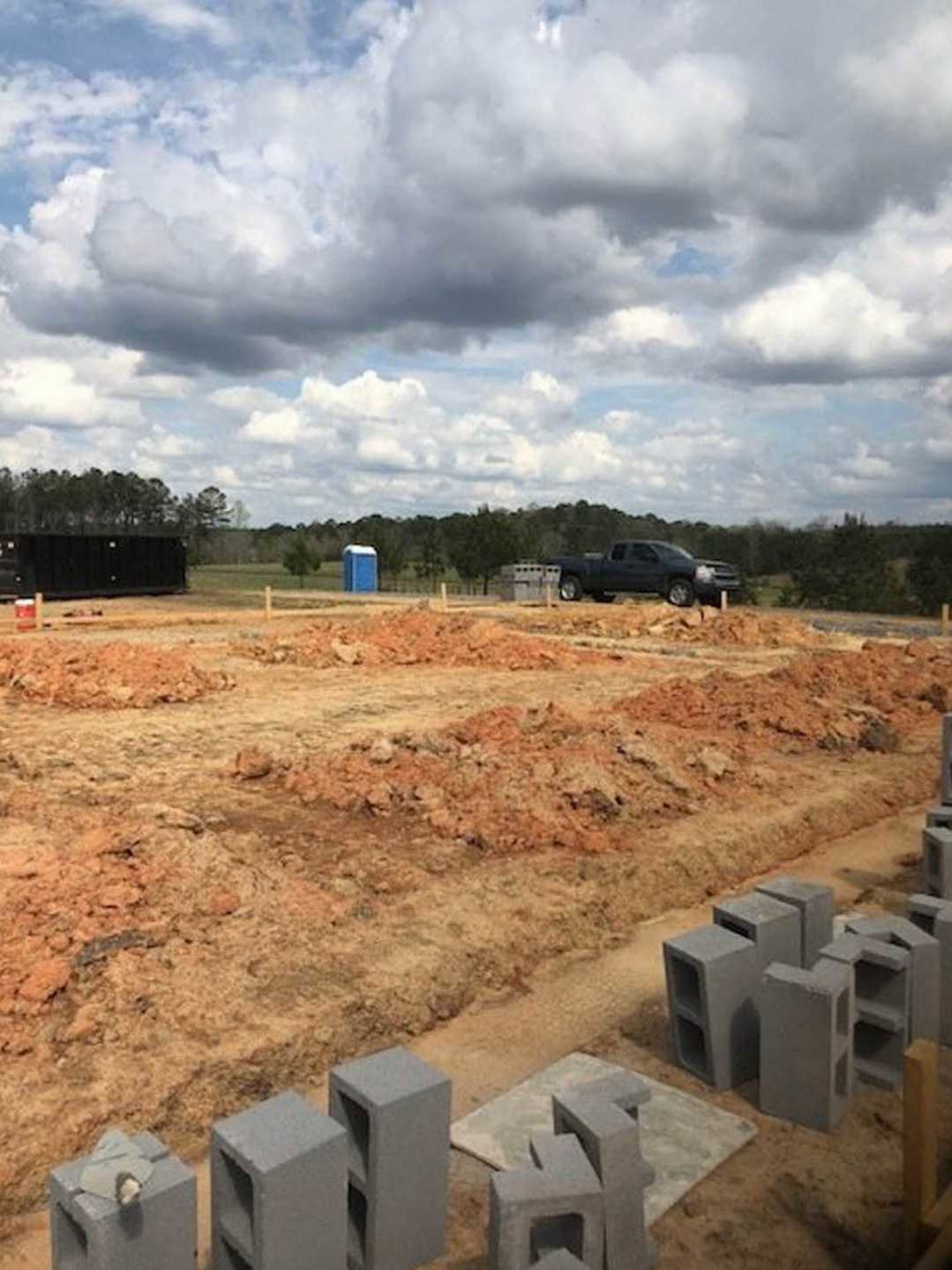 Black pickup truck parked on dirt construction site beside blue shipping container, concrete blocks, and black fence under cloudy sky.
