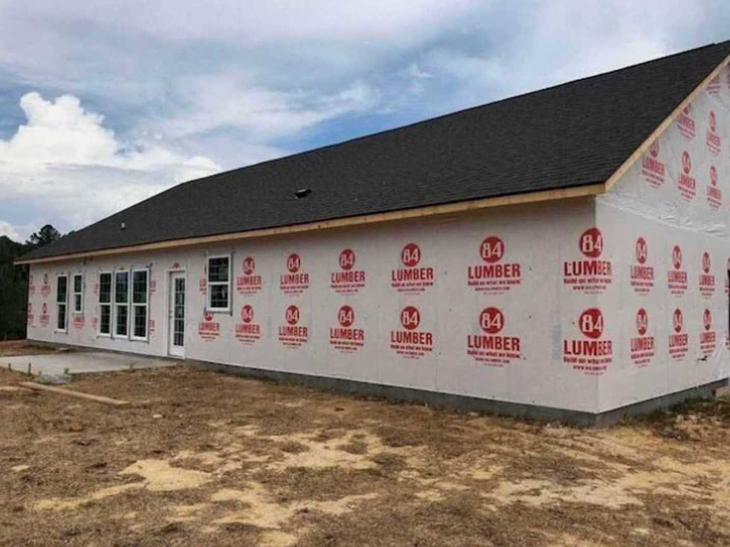 Modern two-story house with white siding, large windows, and red and white logo signs mounted on the exterior wall; dirt and grass landscaping in the foreground under a partly