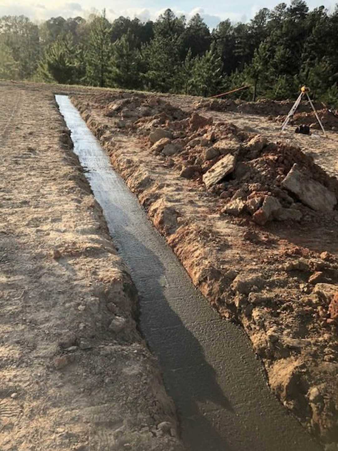 Water-filled drainage ditch running through a dirt field bordered by scattered plants and trees under an open sky