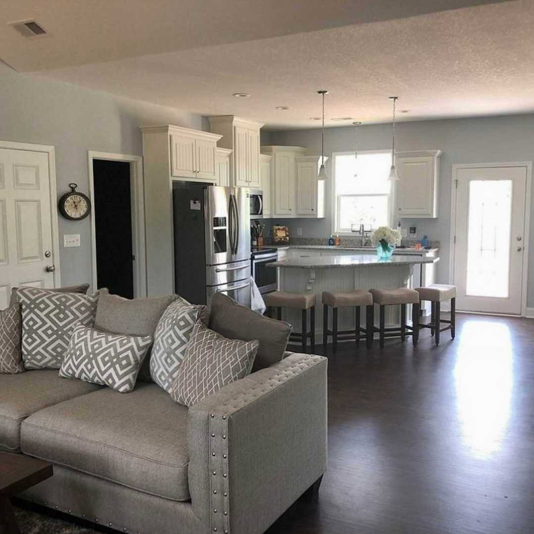 Open concept living room with light gray couch and patterned pillows, adjacent to modern kitchen featuring stainless steel refrigerator, white cabinetry, and wall clock; sunlight