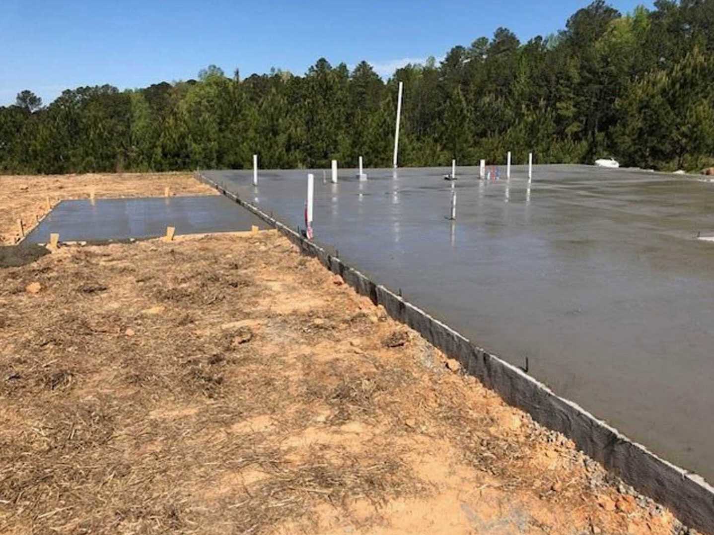 Concrete foundation with upright and fallen poles on bare ground, surrounded by trees and patches of dry grass near water.