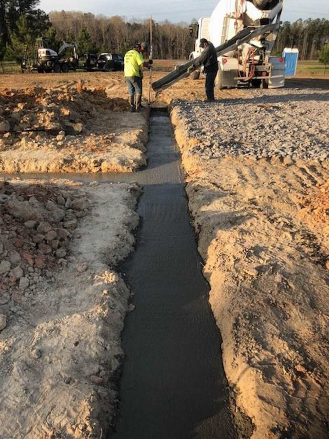 Construction site with several men working on dirt ground, one carrying a cement mixer, another in a yellow shirt, water channel running through soil, car and trees visible in