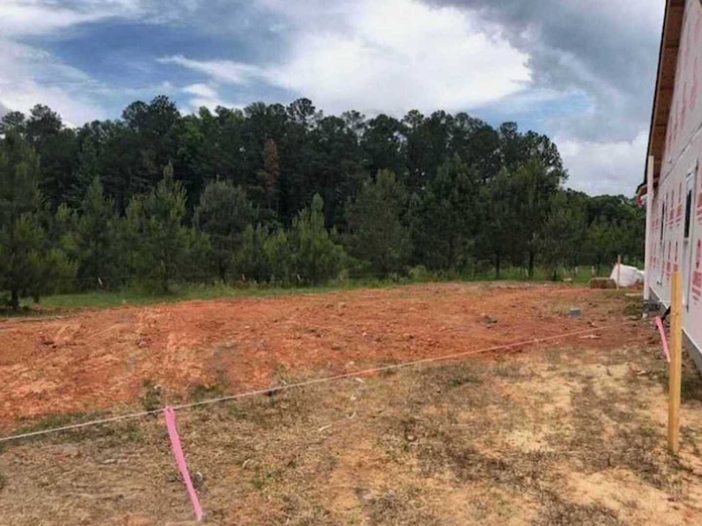 Dirt field with patches of grass, pink line marking ground, trees and forest in background, person holding a sign