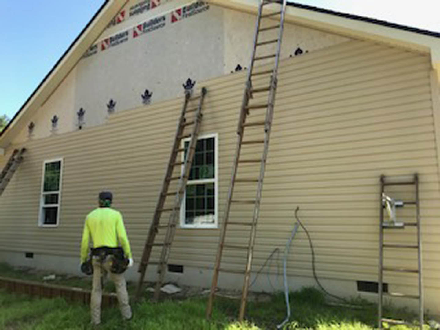 Man in yellow shirt standing beside white house with gray siding, metal ladder leaning against exterior wall, green grass lawn, windows visible, blue sky overhead