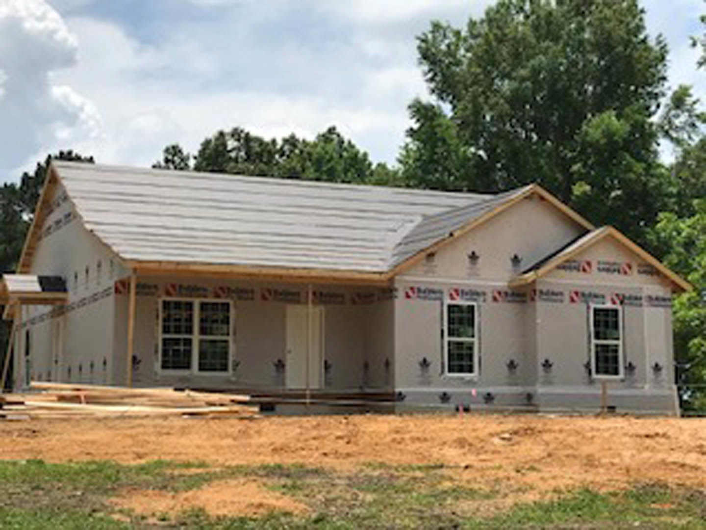 Partially built house with exposed framing, white grid windows, dirt construction site, and mature trees in the background under a cloudy sky