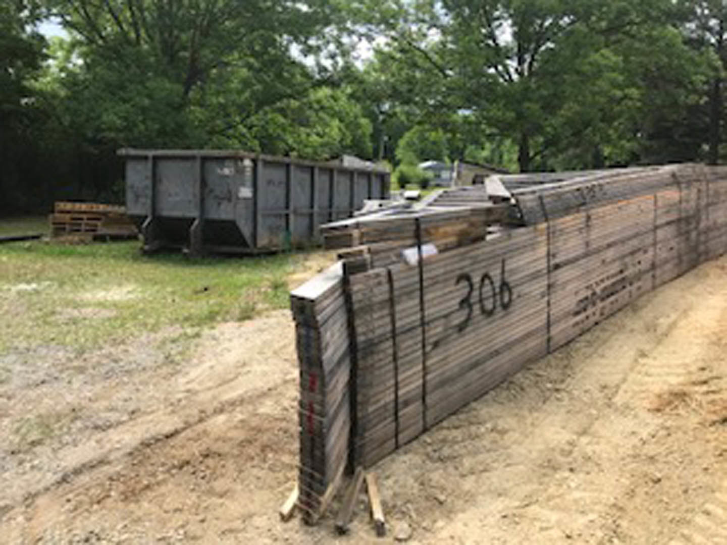 Stack of wood planks on a dirt road beside grass and trees, waste container and building visible in background
