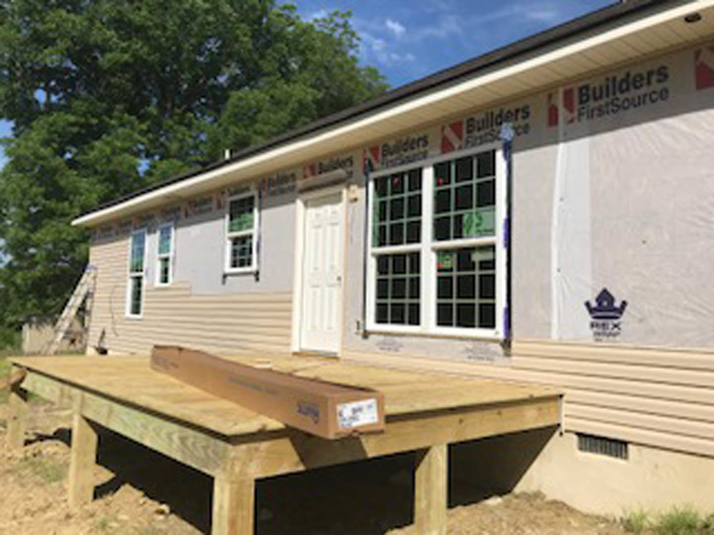 Framed house under construction with exposed lumber, unfinished siding, and a wooden deck; ladder leaning against exterior, windows installed, trees in background
