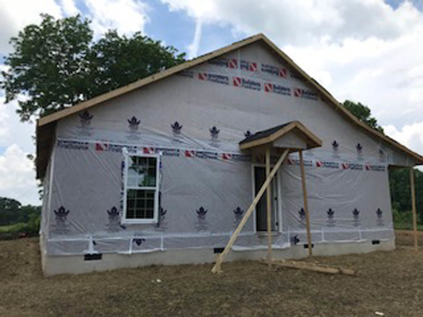 Two-story house under construction with plastic sheeting covering exterior walls, white-framed windows, and a ladder leaning against the unfinished facade