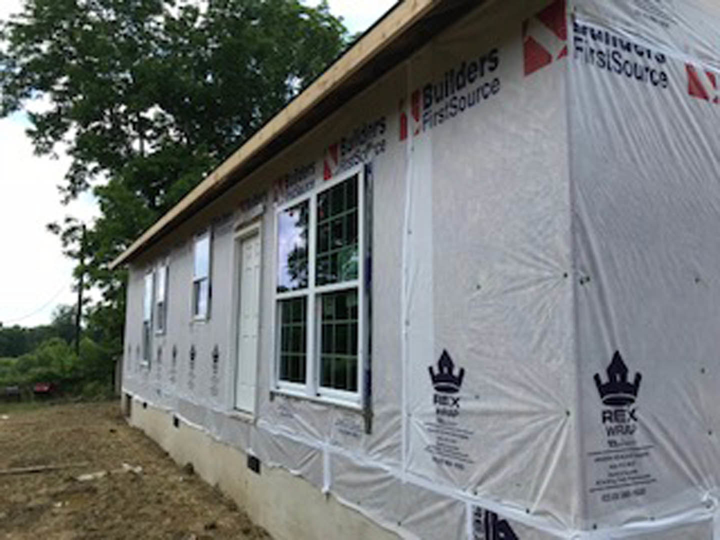 Partially built house with white plastic sheeting, exposed window framing, pitched roof, and mature tree nearby