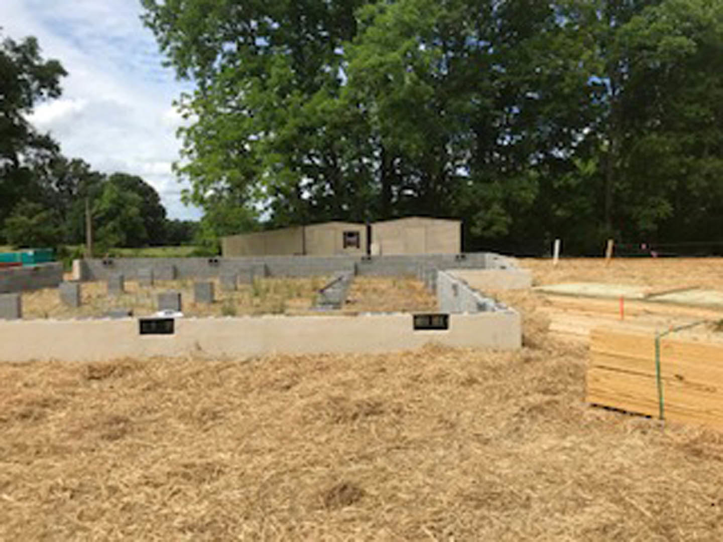 Concrete foundation slab surrounded by dirt and grass, stack of lumber nearby, trees lining the background under a clear sky