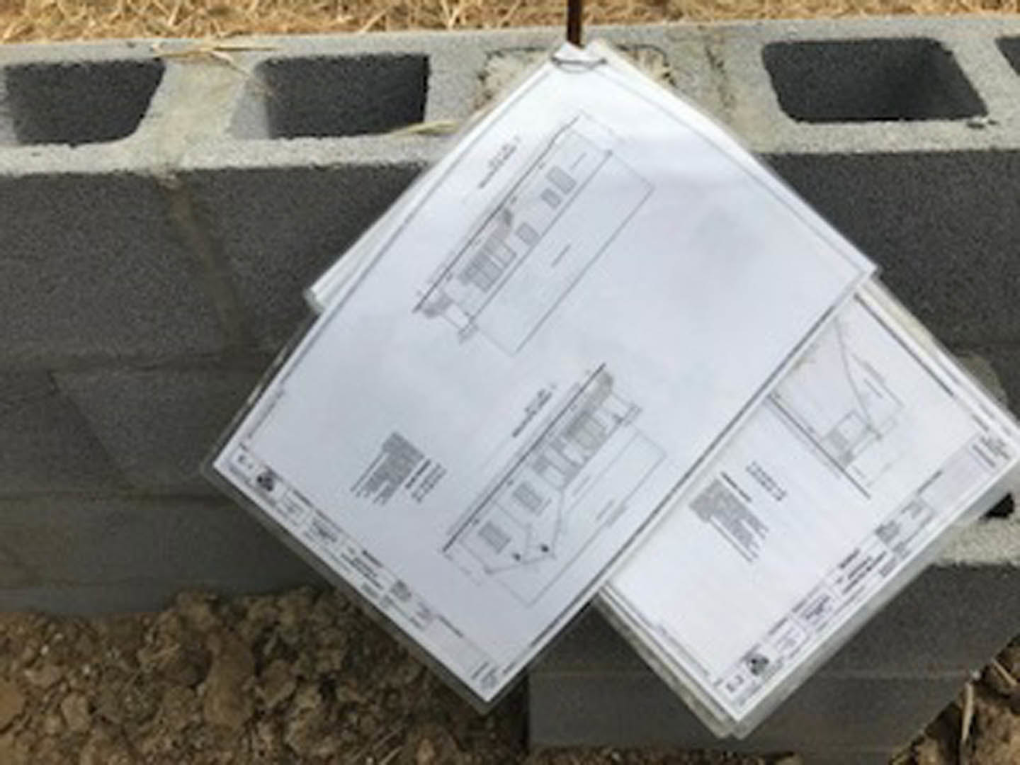 Handwritten paper taped to an exterior brick wall near ground level, with additional papers stacked on a concrete block and a blurry blueprint visible in the background.