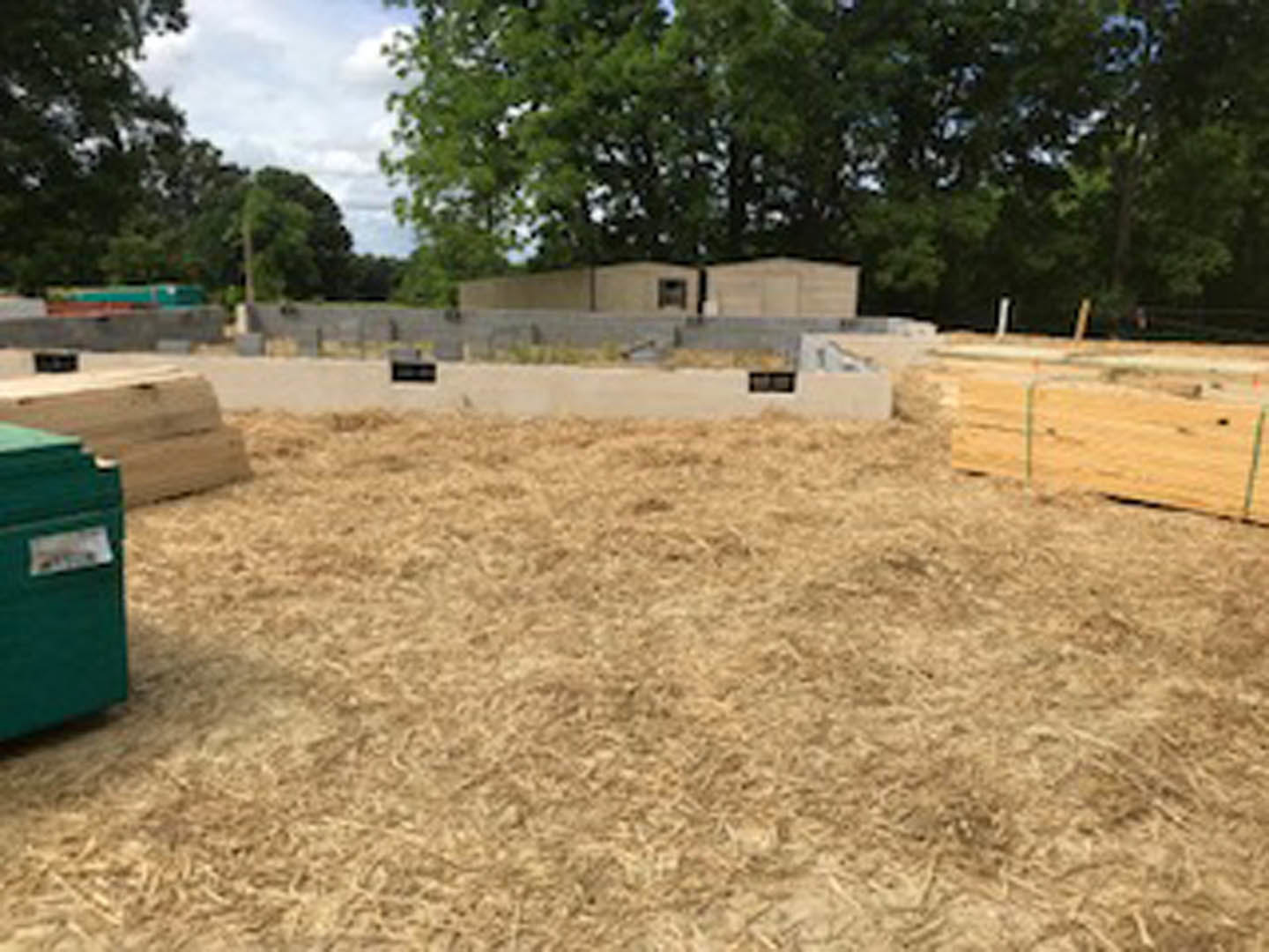 Hay field with scattered wooden pallets, green waste container in foreground, group of trees and concrete wall in background, blue sky overhead