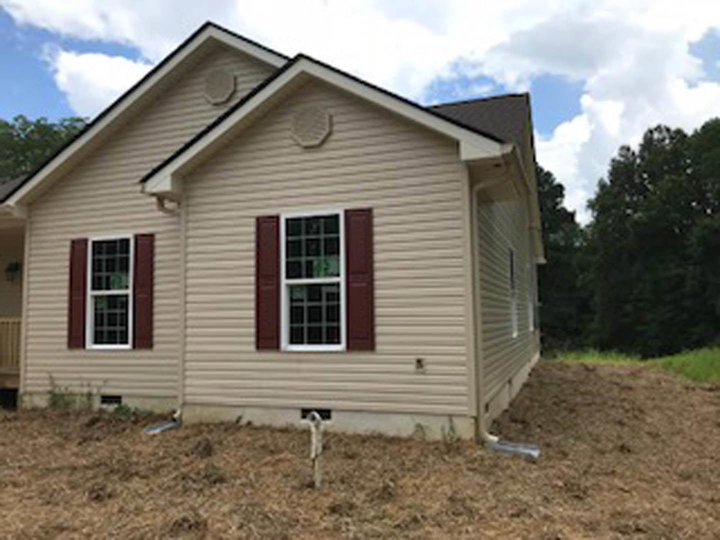 Single-story house with light siding and white-framed windows, surrounded by a dirt yard, wooden fence post, and a dog standing near the entrance under a partly cloudy sky.