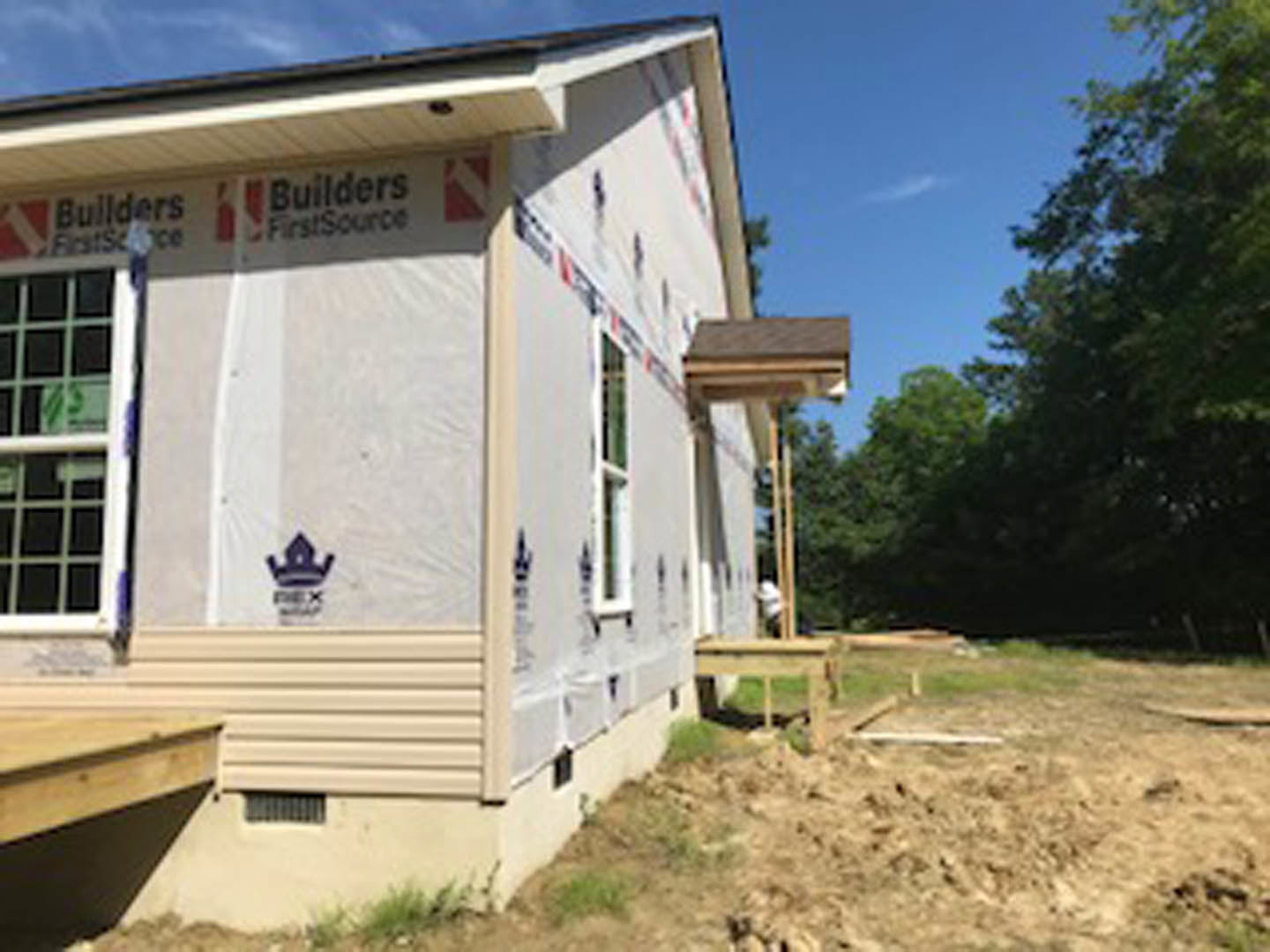 Wood-framed house under construction with exposed deck, surrounded by mature trees, partially covered windows, and plastic sheeting on exterior siding