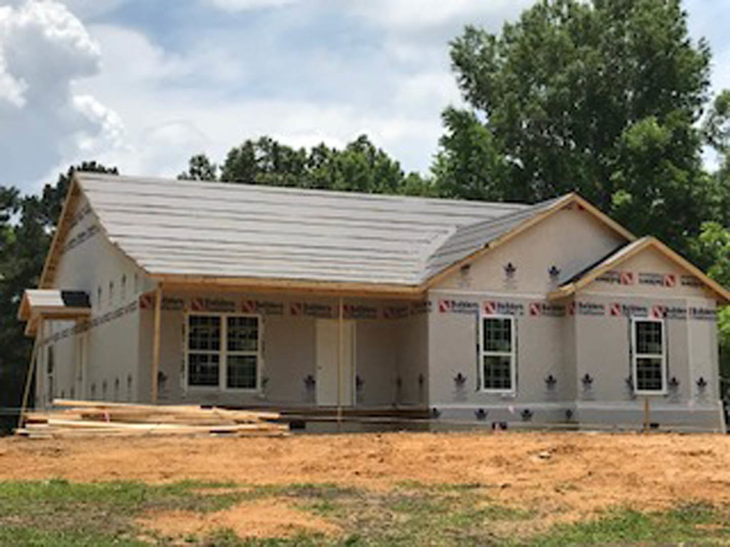Partially built house with exposed wooden framing, white grid windows, and shingled roof, set on grassy lot bordered by trees and a wire fence
