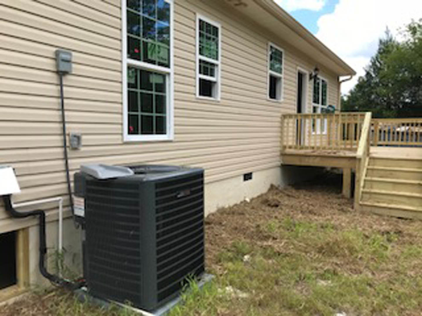 Black heat pump next to wood deck with railing, white-framed window, gray siding, and roof detail
