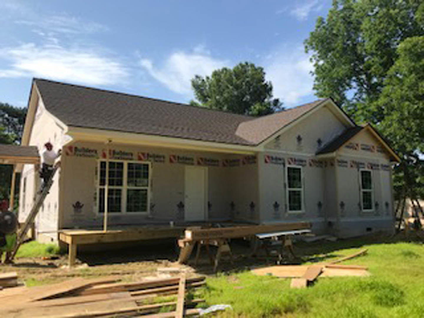 Wood-framed house under construction with exposed beams, unfinished porch, and white-framed window, surrounded by grass and dirt path