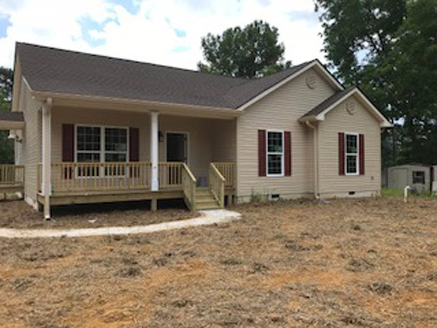 Two-story house with white porch railing, stairs leading to grassy yard, large windows with white frames, surrounded by trees under blue sky