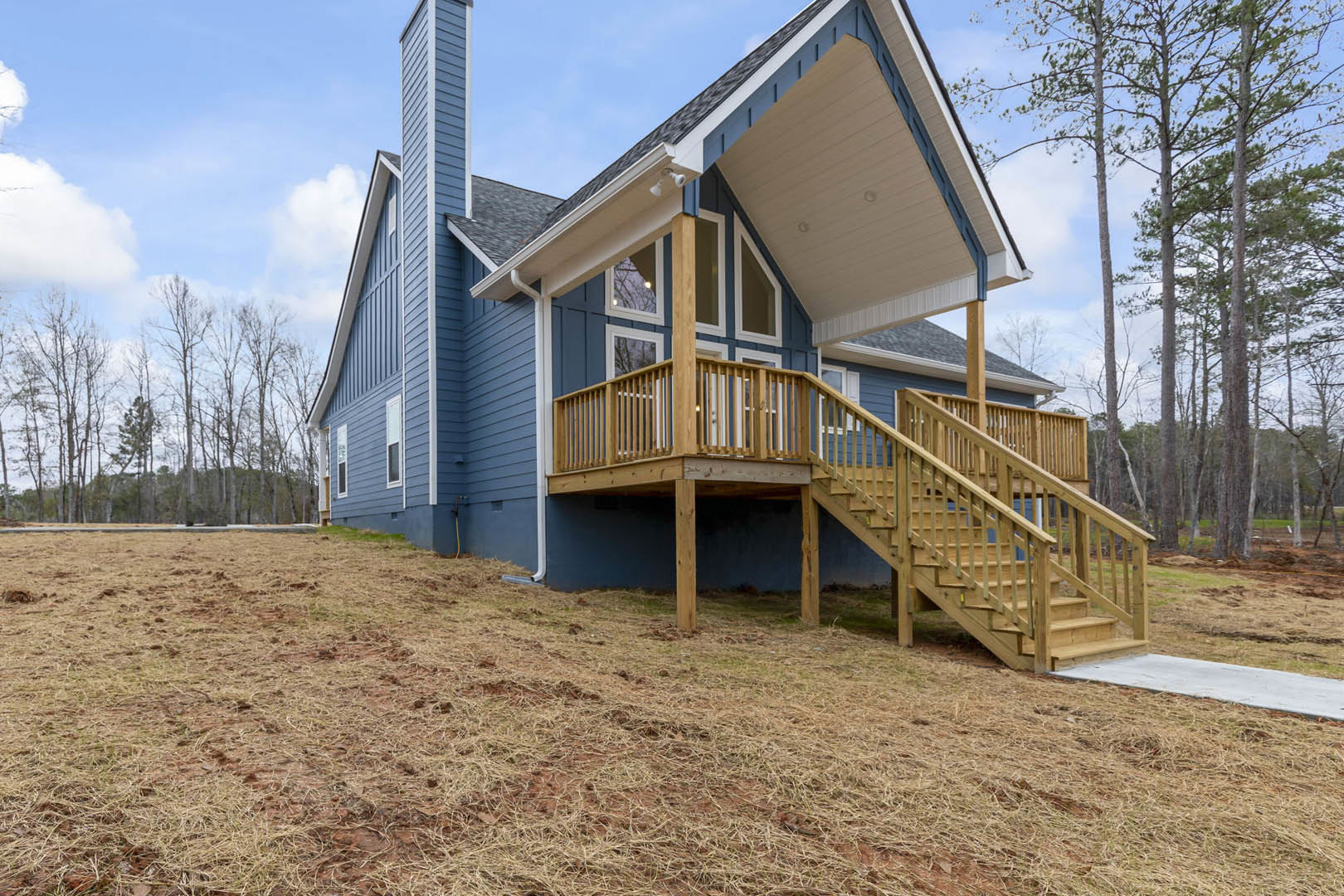 Two-story home with wooden porch and staircase, elevated deck, surrounded by trees, under blue sky with scattered clouds