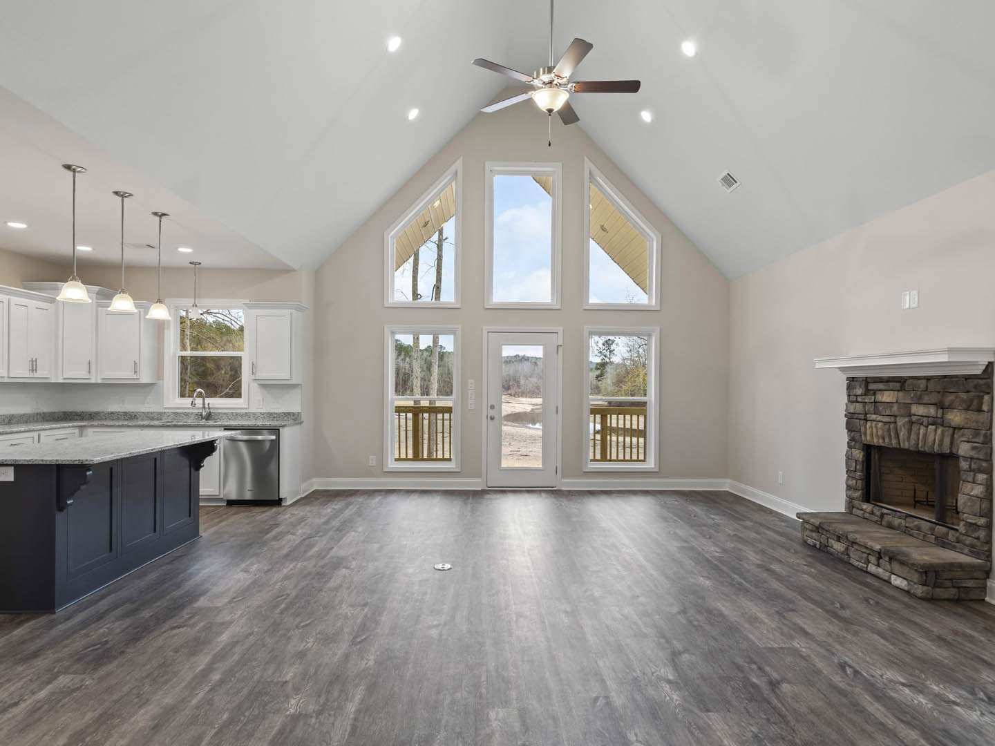 Spacious living room featuring a stone fireplace, ceiling fan with light, wood flooring, white accent furniture, kitchen island with marble countertop, multiple windows, and a door