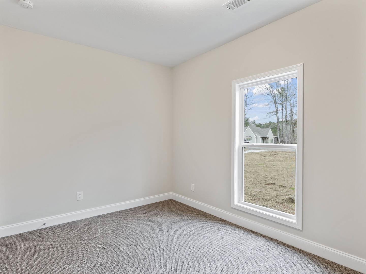 Neutral-toned carpeted room with white walls, large window overlooking green lawn, trees, and neighboring house with parked car visible outside