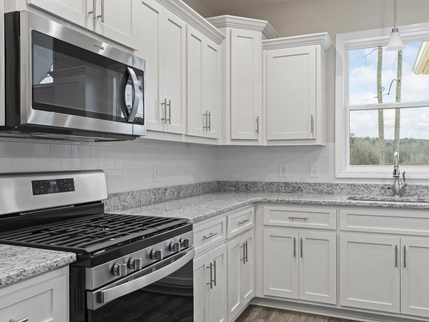 White kitchen cabinets with silver handles, built-in microwave above stove and oven, white countertop, window overlooking trees, white door adjacent to cabinetry.