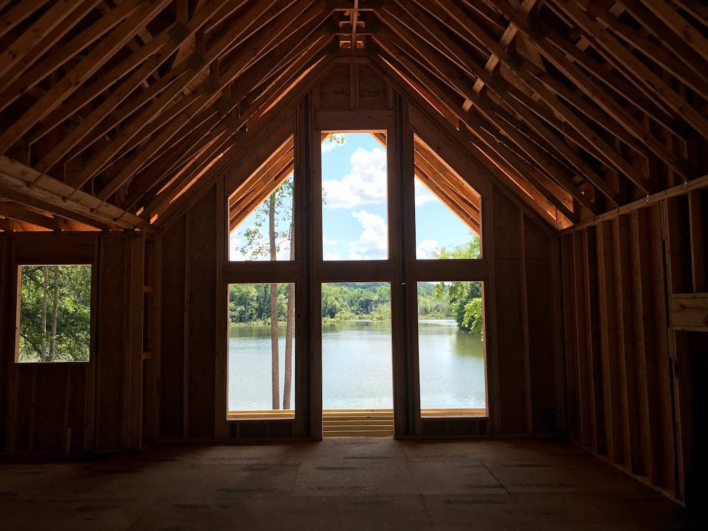 Living room with dark wood flooring, exposed ceiling beams, large windows overlooking a lake and surrounding trees under a blue sky.
