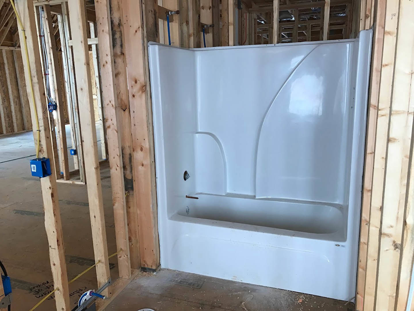 White bathtub set in unfinished bathroom with exposed wooden framing, blue electrical box with yellow wire, ceiling window, and construction materials visible.