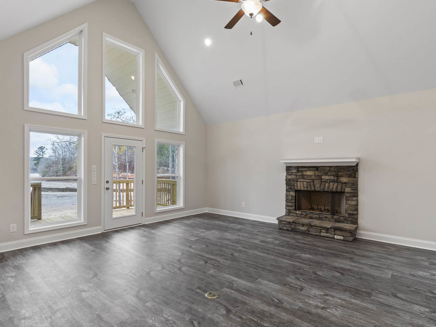 Living room with grey wood flooring, central fireplace featuring a wood bench, ceiling fan with exposed light bulbs, glass door leading to a balcony with railing, white walls and