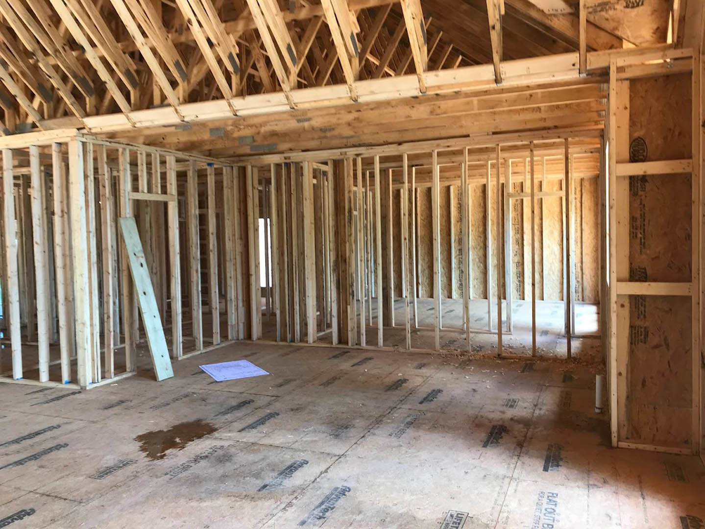 Exposed wooden framing and beams inside a house under construction, with unfinished hardwood floor, insulation visible between studs, and construction markings on the subfloor.