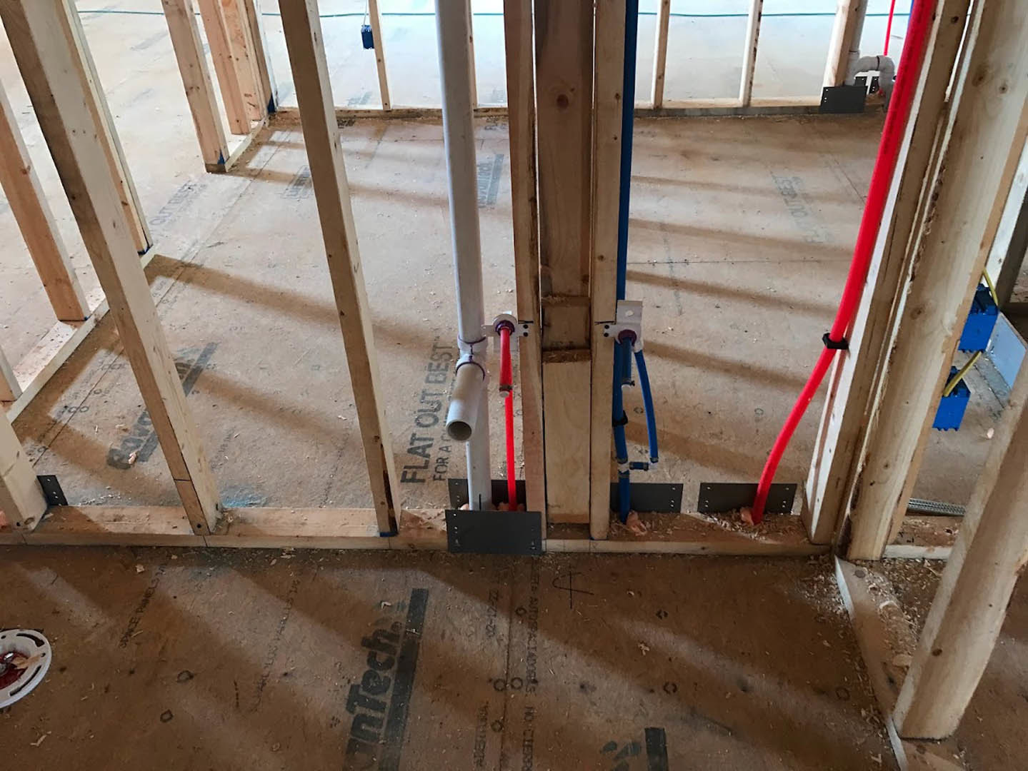 Exposed plumbing pipes and wooden framing inside a partially constructed room, with insulation visible between beams and a metal ladder leaning against the wall.