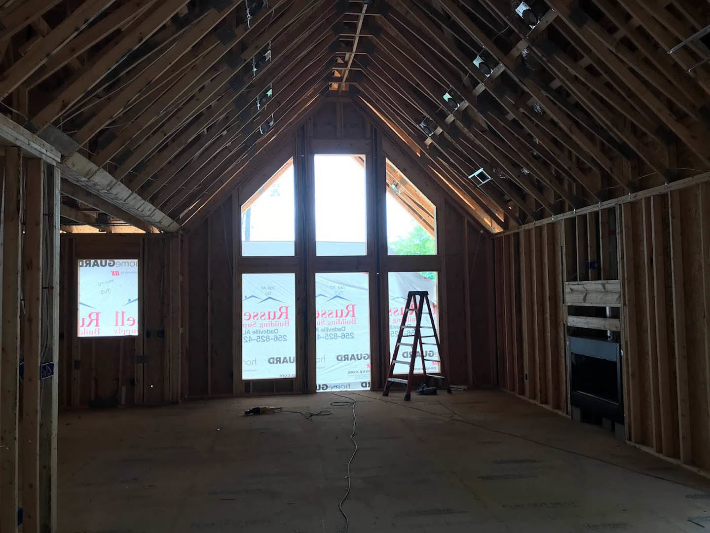 Sunlit attic room with exposed wooden beams, tall windows, and a metal ladder leaning against the wall; light hardwood floors and white walls.