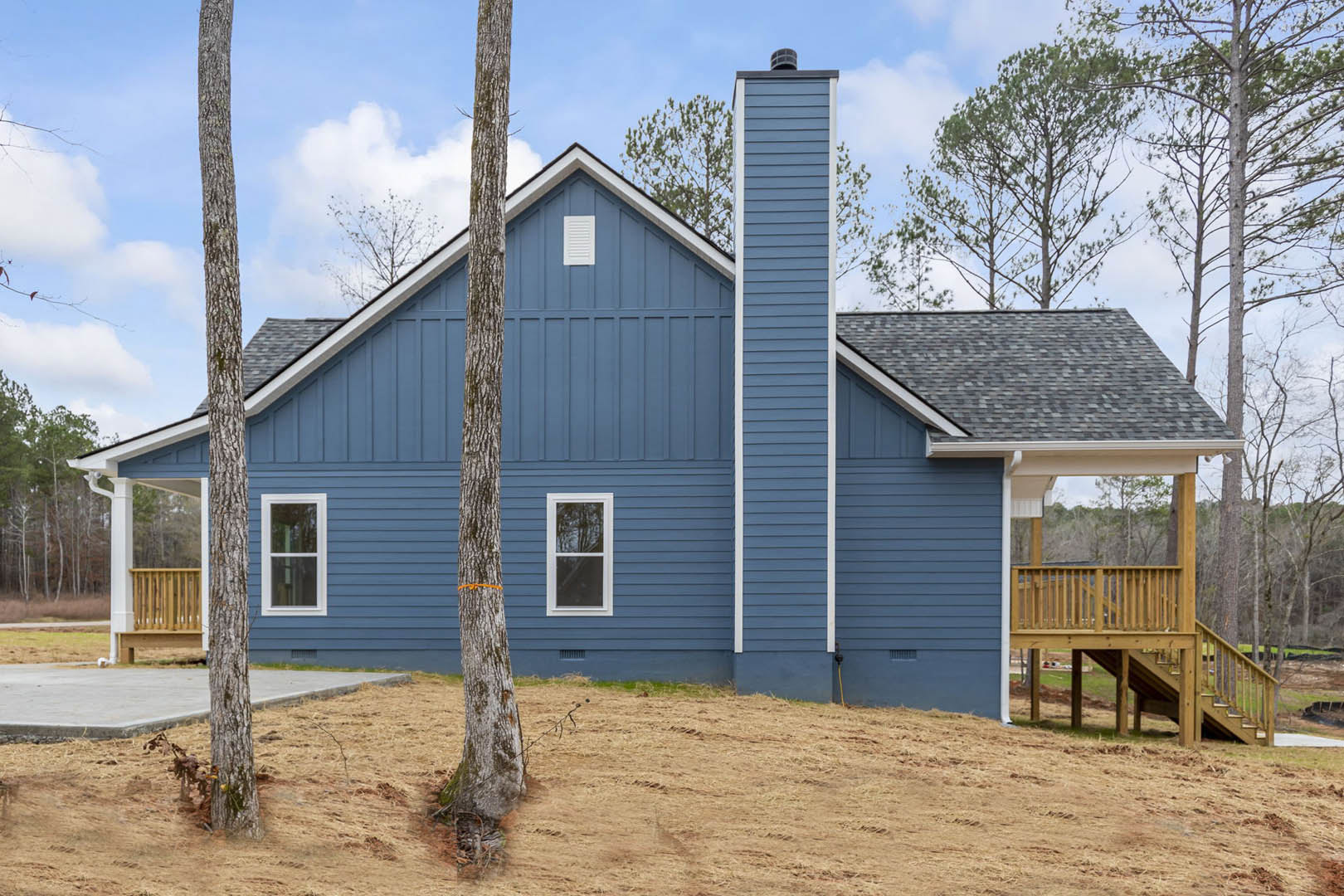 Blue siding exterior with white-framed windows, elevated deck, brick chimney, surrounded by mature trees under a partly cloudy sky
