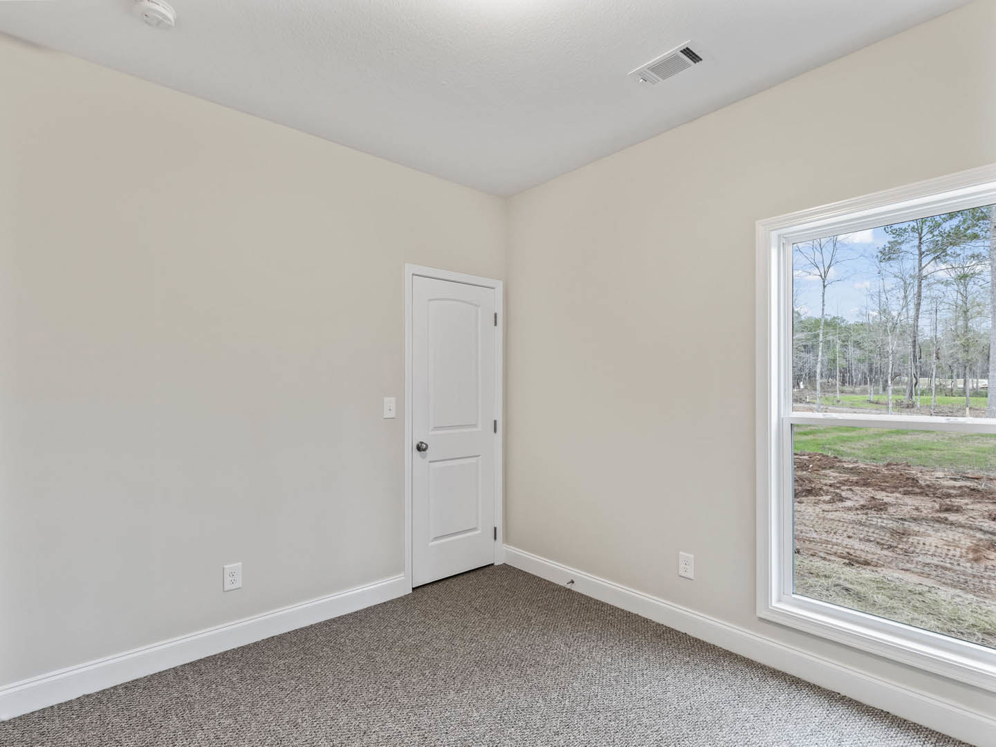 Carpeted room with white door featuring silver knob, large window overlooking dirt field and trees, white plaster walls