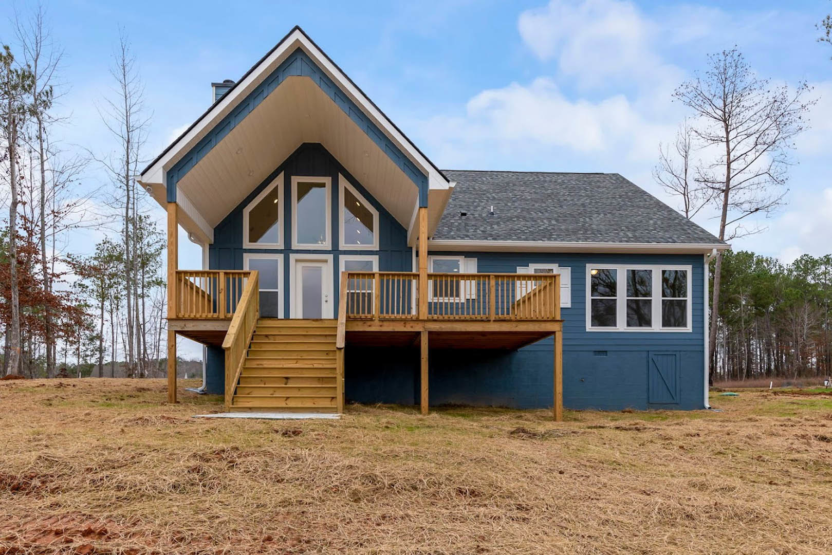 Two-story home with light siding, wooden deck and railing, covered front porch, stairs leading to lawn, large windows reflecting nearby trees, cloudy sky overhead