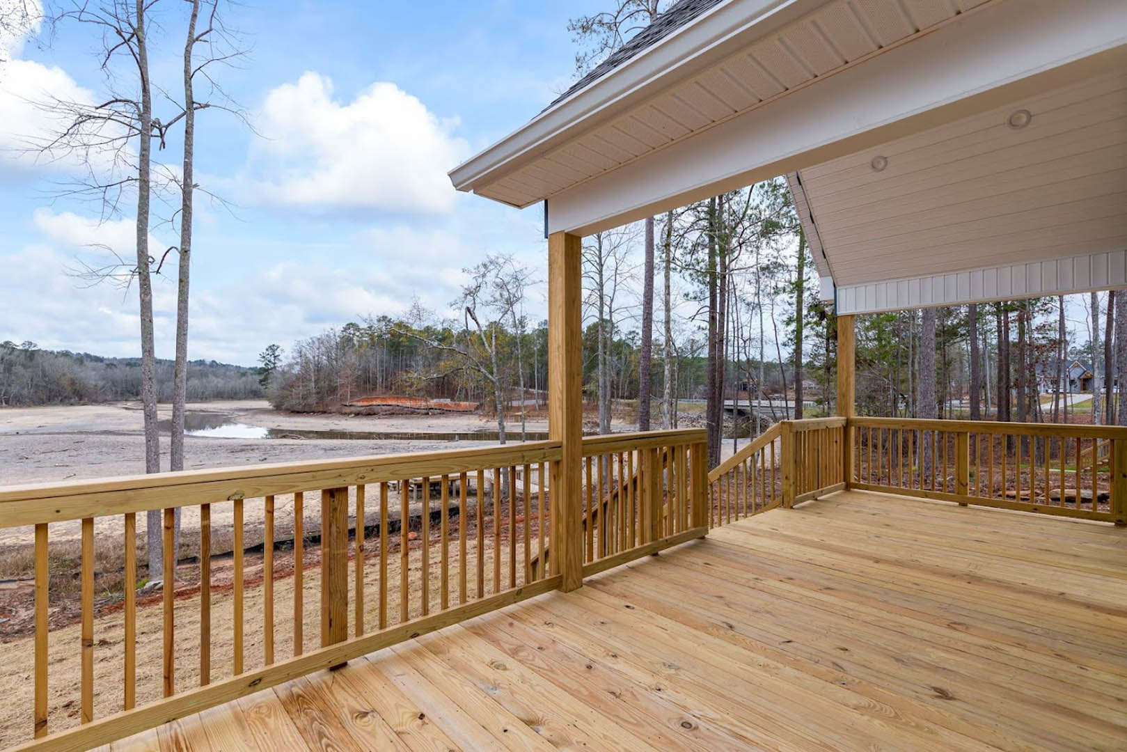Wooden deck with railing overlooking a lake and tall trees, partially shaded by roof, under a cloudy sky
