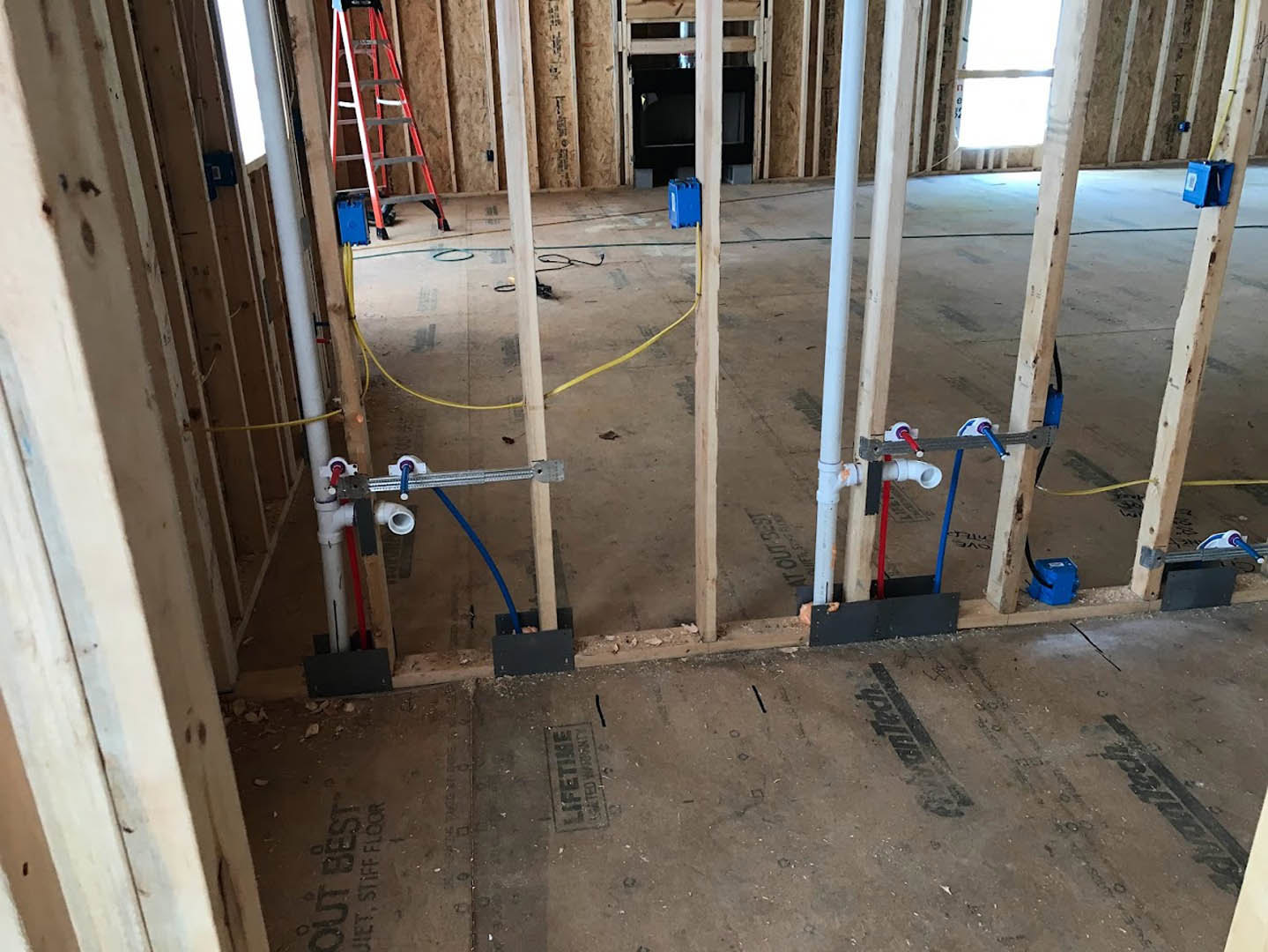 Utility room under construction with exposed pipes and wires, a metal ladder, and a blue labeled box on the concrete floor.