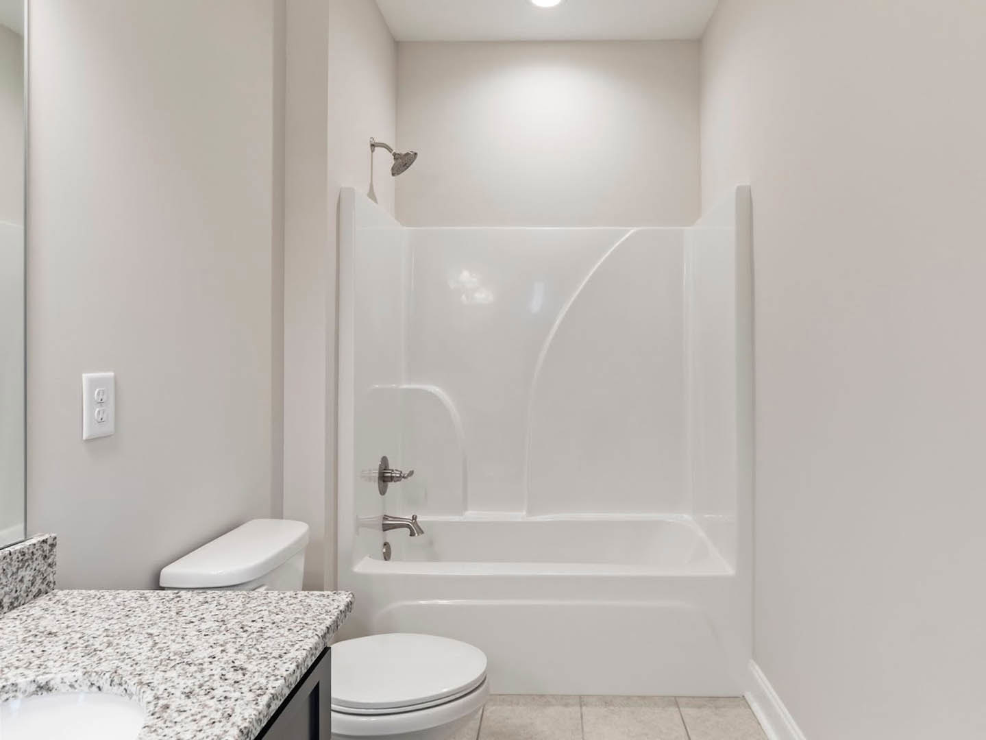 Modern bathroom featuring a white bathtub with shower, white toilet with lid up, light-colored tile walls, built-in sink, and a close-up of a stone countertop and electrical