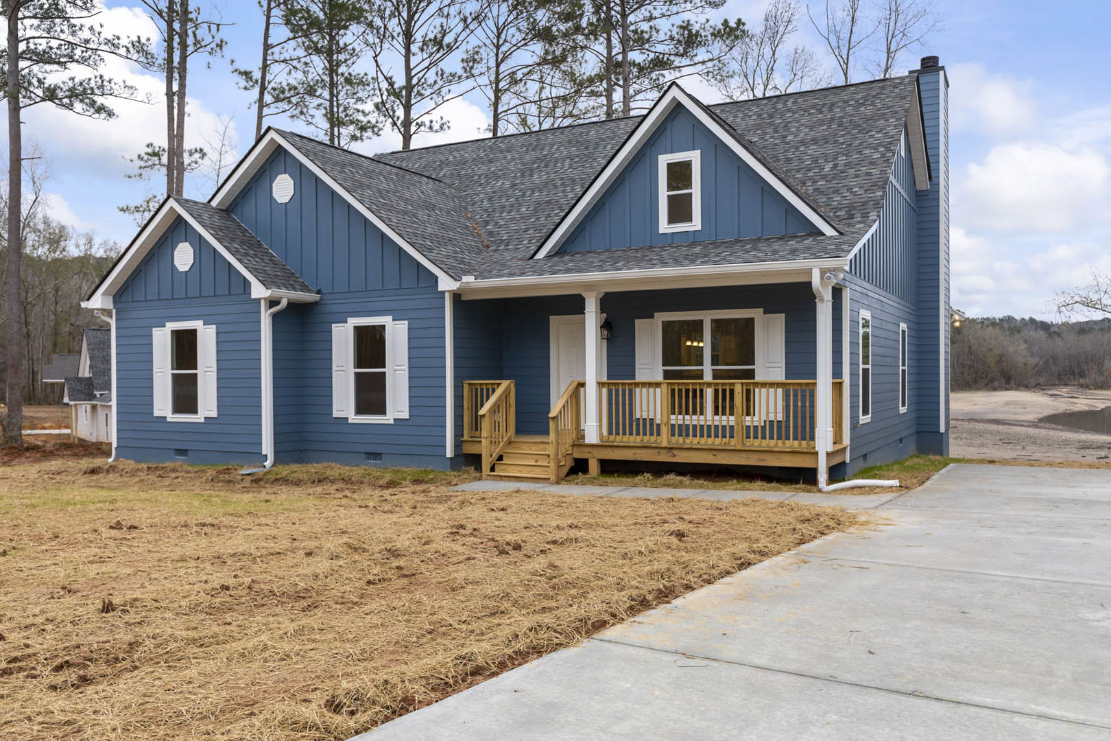 Blue siding house with white trim, grey roof, covered porch with wooden railing, concrete driveway, green lawn, mature trees, and cloudy sky.