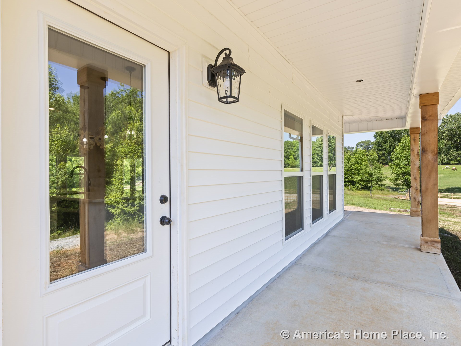 Covered front porch with white horizontal siding, glass-paneled entry door, wood support columns, concrete flooring, triple window unit, black outdoor wall lantern, and white trim