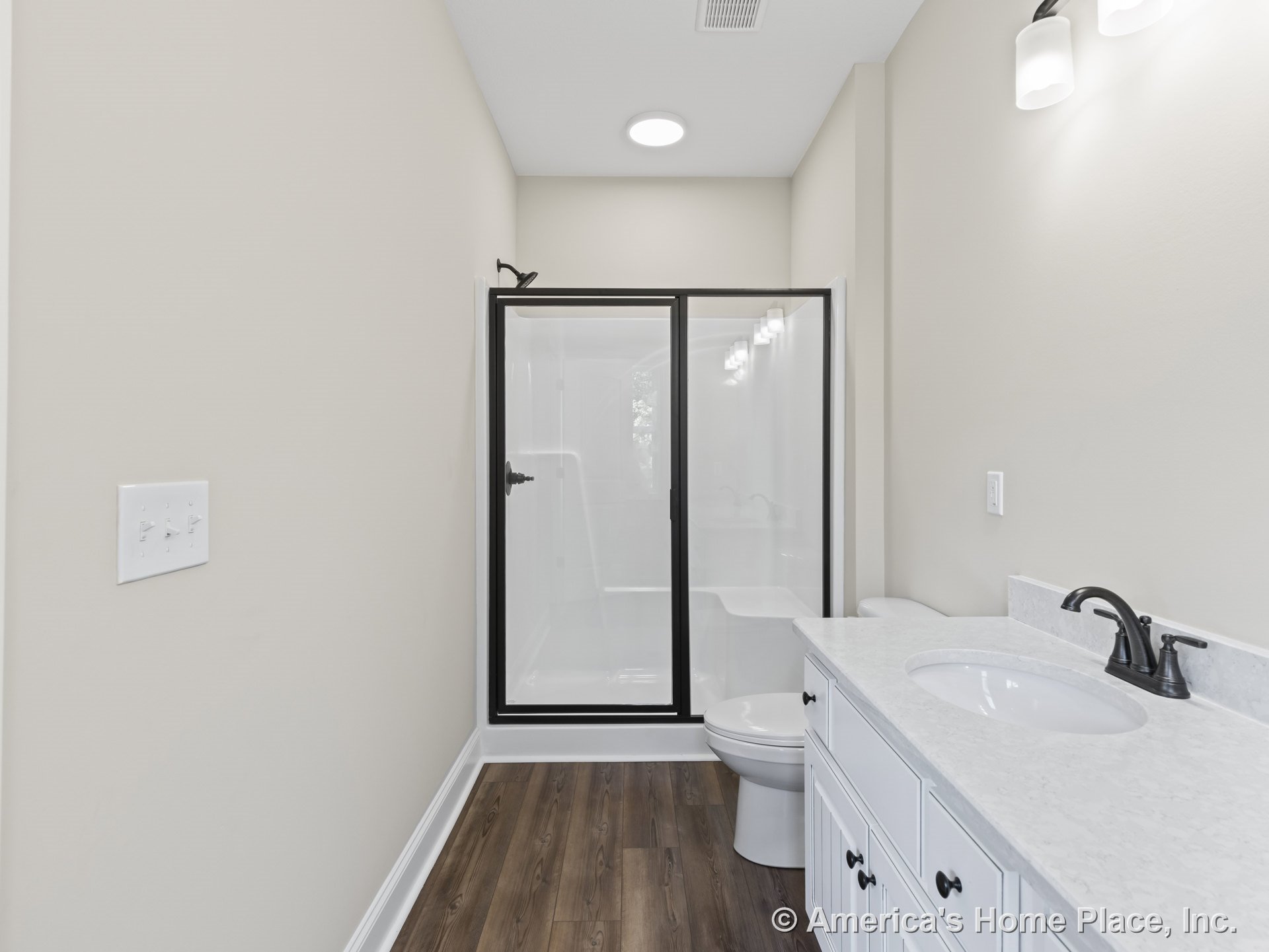 Glass-enclosed shower with matte black fixtures, white vanity cabinetry topped with quartz countertops, wood-look flooring, wall-mounted lighting, and neutral walls with crisp