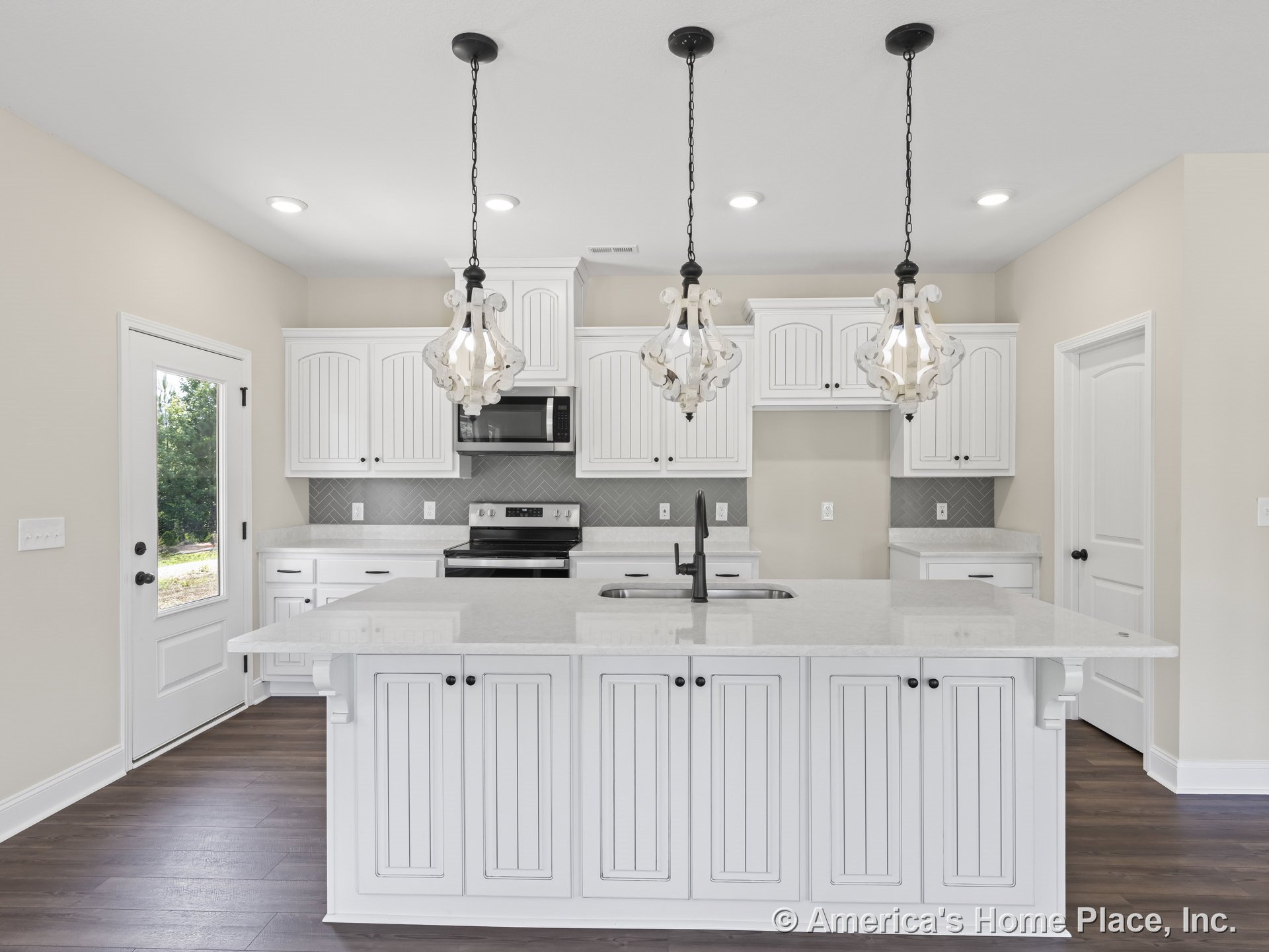 White shaker cabinets and quartz countertops surround a spacious kitchen island with built-in sink, topped by three decorative pendant lights; herringbone tile backsplash and