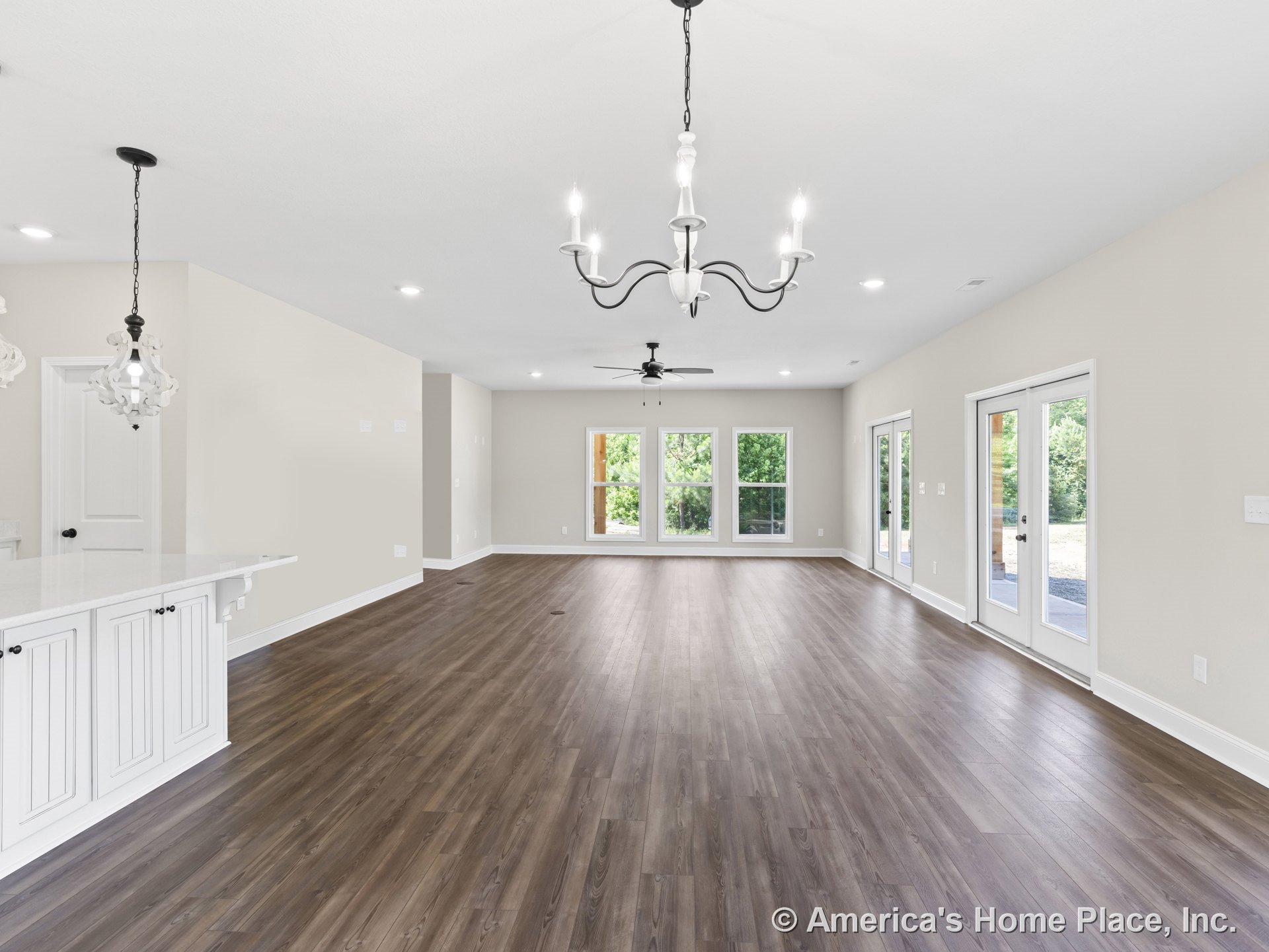 Open concept living area with wide plank wood floors, multiple large windows, French doors to patio, white kitchen cabinetry, ceiling-mounted pendant lights, chandelier, ceiling
