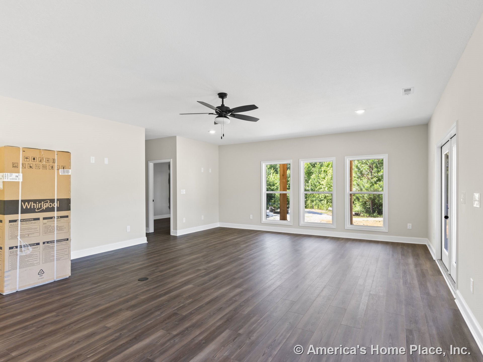 Spacious living room with three-pane windows, dark wood-look flooring, white baseboards, neutral wall paint, ceiling fan with light, and glass exterior door allowing natural light.