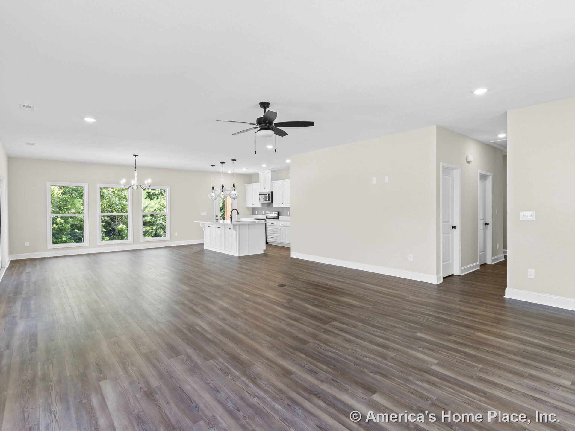 Open-concept living area with wide plank wood flooring, three large windows, modern kitchen featuring white cabinetry, pendant lighting above island, ceiling fan with lights