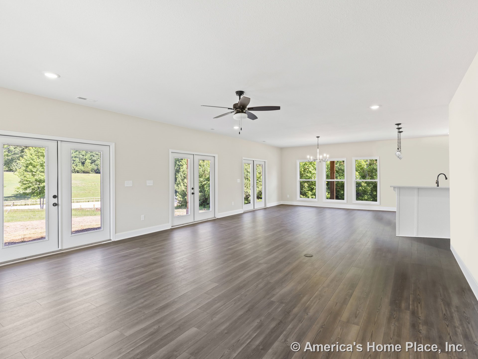 Open-concept living area with wide plank wood-look flooring, multiple large picture windows, French doors, recessed ceiling lights, ceiling fan, chandelier, and adjoining kitchen