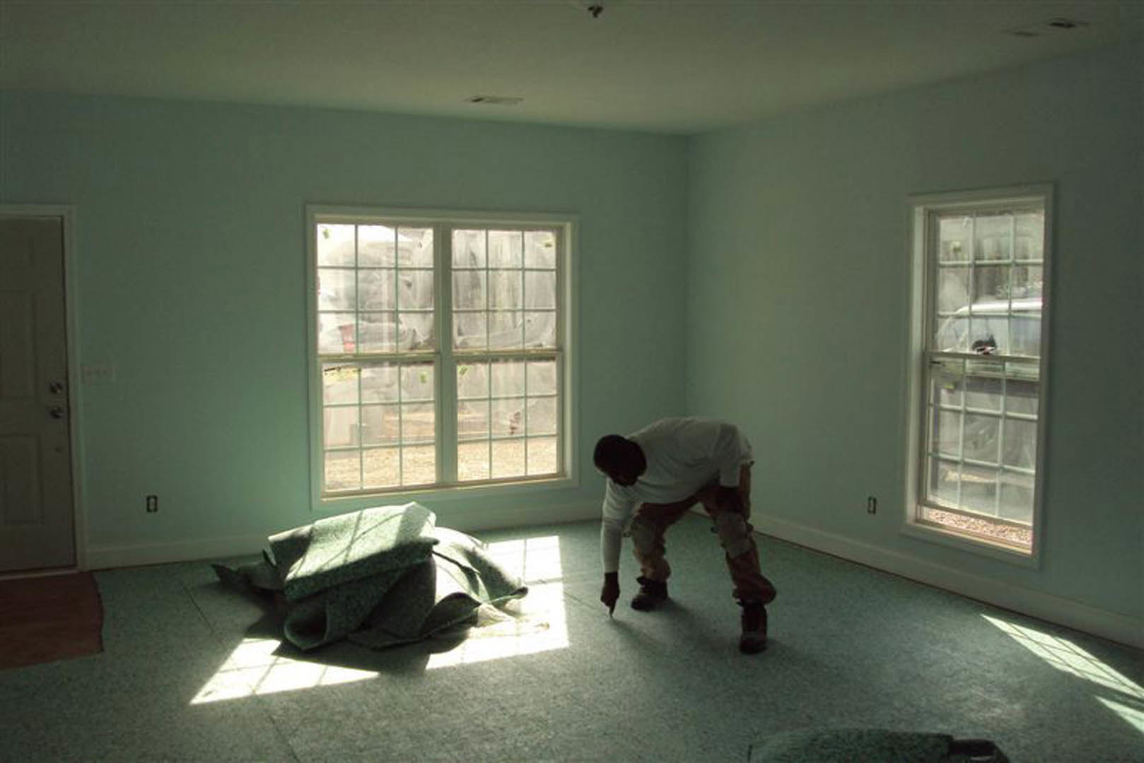 Man in white shirt cleaning a room with light wood flooring, white plaster walls, large window with white frame, folded blanket draped over chair
