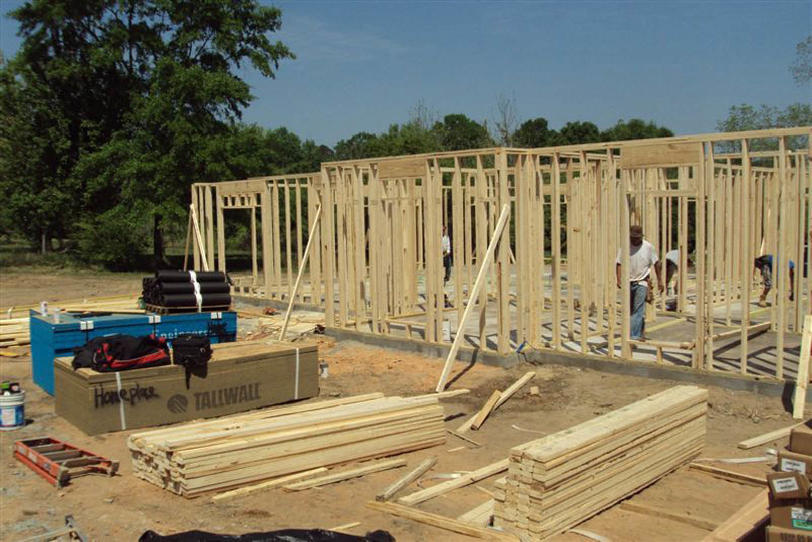 Framed house under construction with workers on site, exposed foundation, stacks of black pipes, scattered building materials, trees and blue sky in background