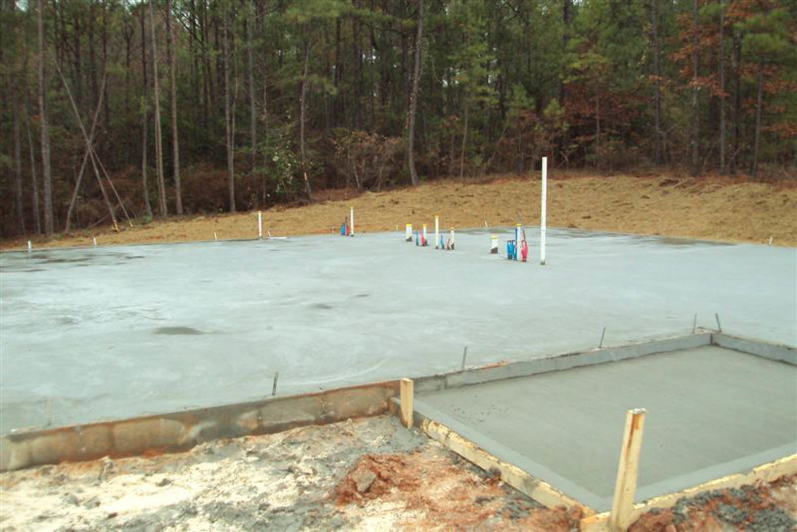 Concrete foundation with upright wooden poles surrounded by dense forest and natural ground.