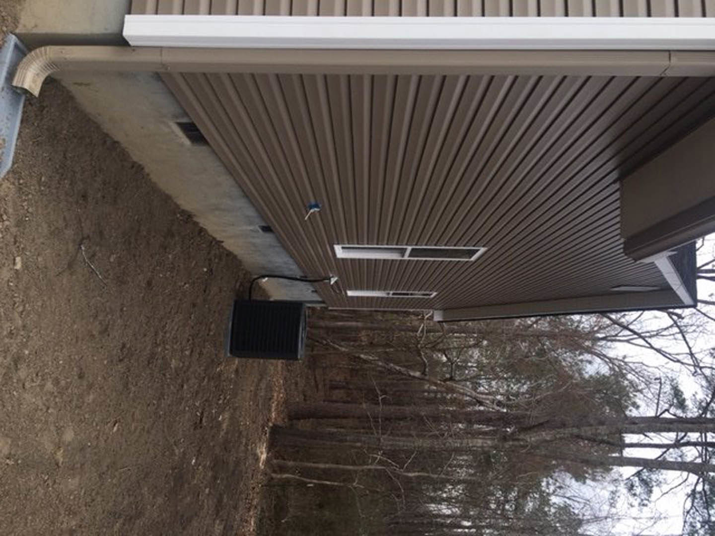 Brown metal exterior wall with white trim, black square vent, and metal door set in dirt foreground; forest visible through window.